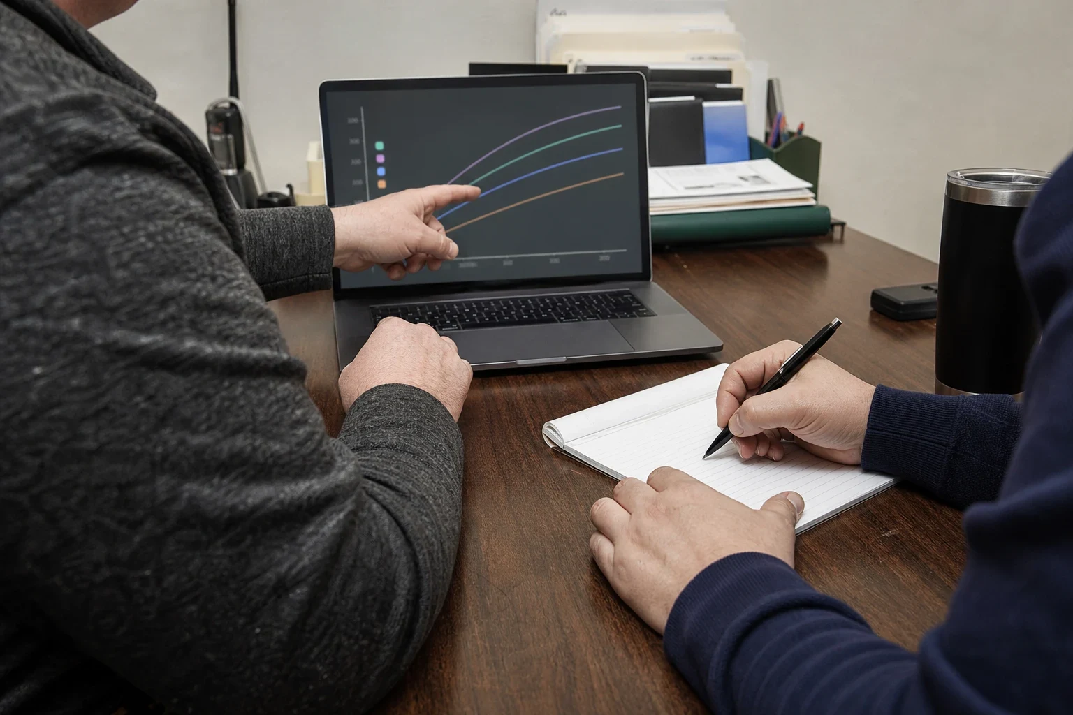 Over-the-shoulder view of a man pointing at a growth chart on a laptop while another person takes notes on a legal pad at a wooden office desk
