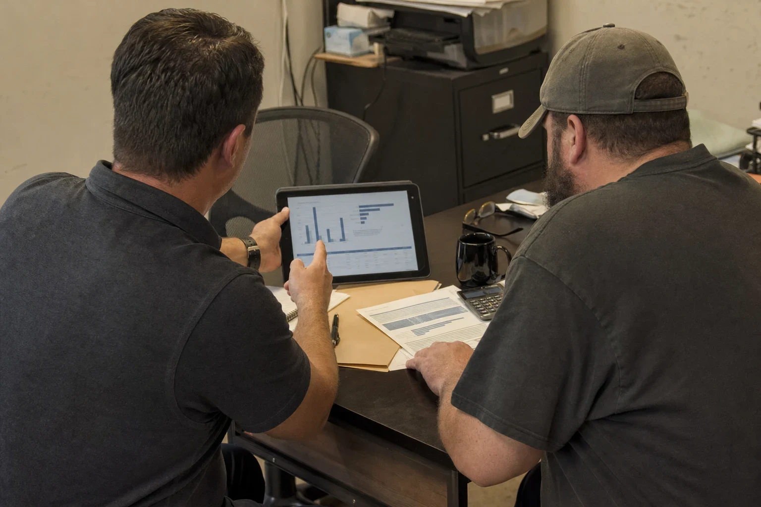Two men at a desk in a small back office, one holding a tablet showing bar charts while the other reviews a printed sheet beside a calculator and coffee mug