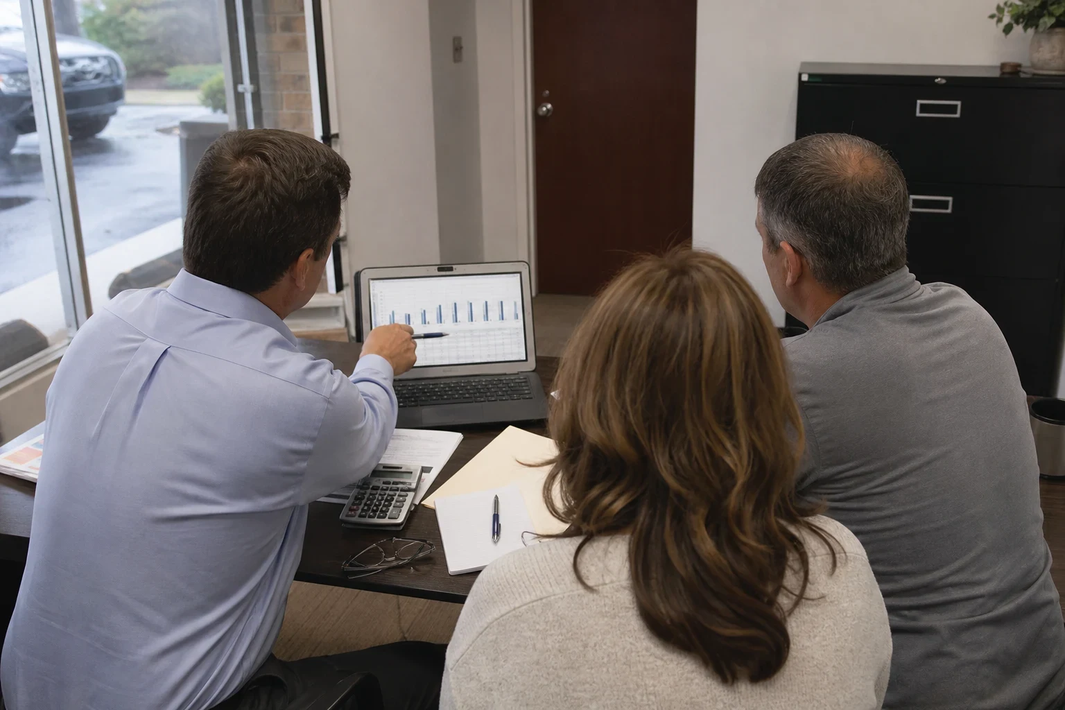 Agent in a light blue shirt points to a bar chart on a laptop while a couple looks on at a small table in a storefront office with a window and filing cabinet.