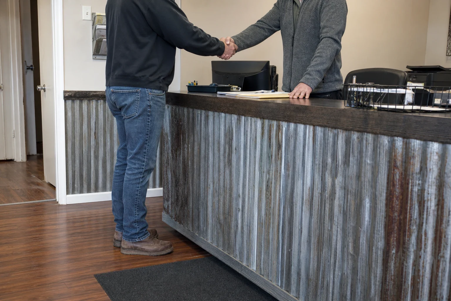 Two men shake hands across a rustic corrugated-metal shop counter with a manila folder, papers and a computer monitor on the countertop.